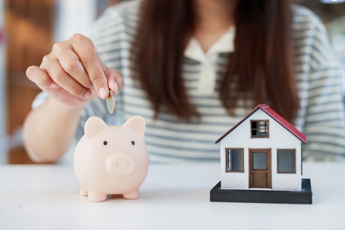 Woman saving coins in a piggy bank beside a house model, representing financial planning for home ownership